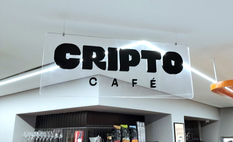 Placa de sinalização interna suspensa com a inscrição "CRIPTO CAFÉ". Base transparente em acrílico cristal de 8mm com letras em acrílico preto de 6mm, instalada no teto através de cabos de aço.
