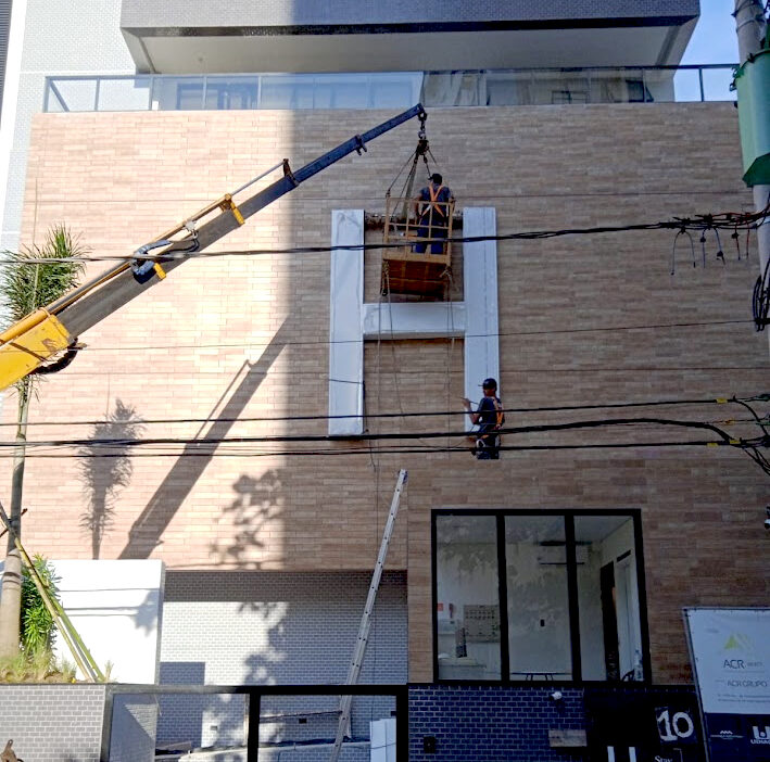 Equipe técnica da Jonatan Mattos Soluções Visuais operando em um cesto de guindaste para realizar a instalação de uma letra caixa gigante em formato de 'H' na fachada de tijolos de um edifício comercial.